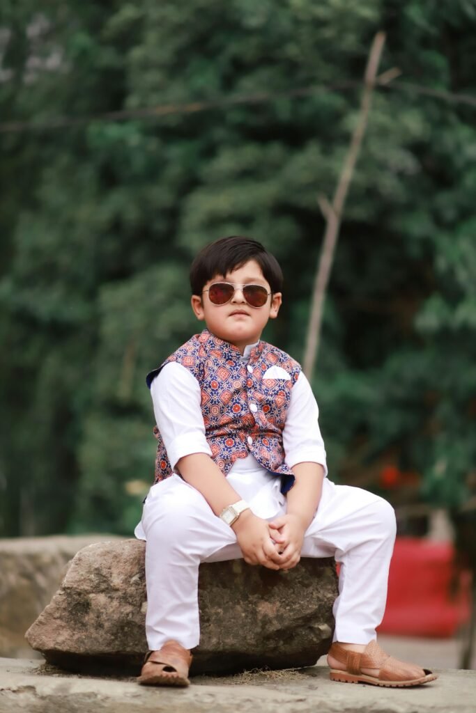 Young boy sitting outdoors wearing sunglasses and traditional attire, exuding confidence.