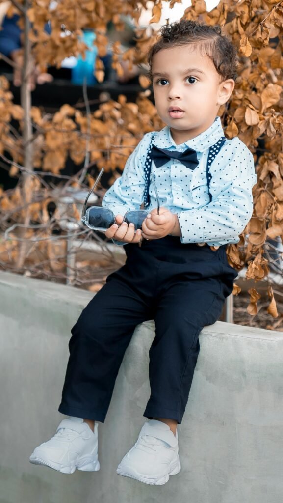 Charming toddler in bow tie and suspenders sitting outdoors holding sunglasses.