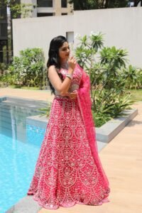 An elegant woman in a vibrant pink lehenga poses gracefully by a poolside during a sunny day.